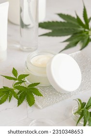 Opened white cream jar with a lid near green cannabis sativa leaves on a marble table. Cosmetic Mockup, Copy space. Organic skincare beauty product. Eco friendly body or hand cream with hemp - Shutterstock ID 2310348653