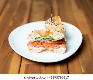 An open-faced bagel sandwich with layers of smoked salmon, cream cheese, avocado slices, tomato, and red onion, sprinkled with sesame seeds on a white plate - Powered by Shutterstock