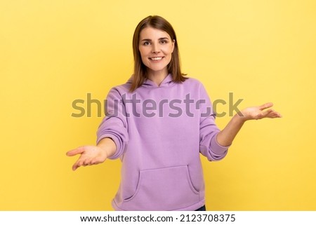 Openhearted generous woman outstretching hands looking at camera with kind smile, greeting and regaling, happy glad to see you, wearing purple hoodie. Indoor studio shot isolated on yellow background.