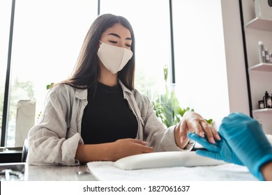 Opening Of Beauty Studio After Lockdown. Asian Woman Client In Protective Mask Looks At Master In Rubber Gloves, Doing Manicure And Files Nails In Salon On Window Background, Cropped, Free Space