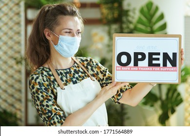 Opening Small Business After Coronavirus Pandemic. Stylish Small Business Owner Woman In Apron With Medical Mask Showing Welcome Sign.