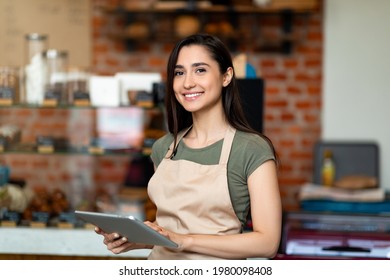 Opening small business. Happy arab woman in apron near bar counter holding digital tablet and looking at camera, waiting for clients in modern loft cafe