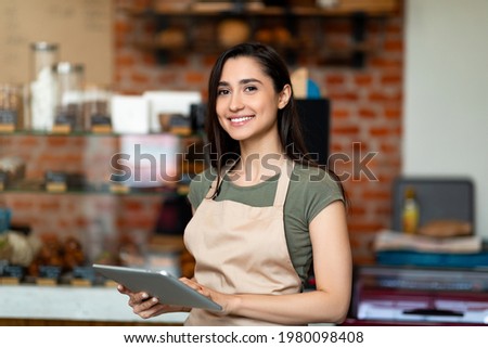 Opening small business. Happy arab woman in apron near bar counter holding digital tablet and looking at camera, waiting for clients in modern loft cafe