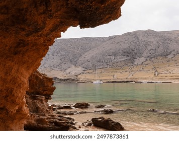 Opening of unique rock formation unfolds an idyllic coastal vista in Vela Luka bay on island Otok Krk, Primorje-Gorski Kotar, Croatia. Calm turquoise sea, a distant sailboat, and mountainous backdrop - Powered by Shutterstock