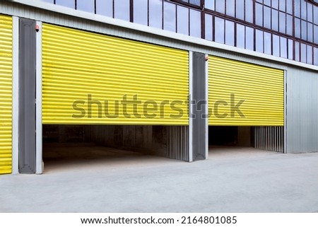 Opening yellow iron shutter door of garage and industrial building warehouse exterior facade with grey concrete road, side view nobody.