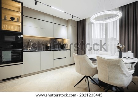 Open-plan kitchen area featuring handleless beige cabinets, dark marble apron, built-in appliances, glass display case, light herringbone parquet flooring, and circular LED ring pendant light above dining table.