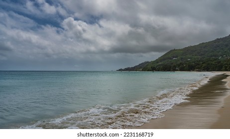 The openwork foam of the waves spreads over the beach. Wet sand glistens. The turquoise ocean is calm. A green hill against the sky and clouds. Seychelles. Mahe. Beau Vallon - Shutterstock ID 2231168719