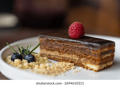 Opera cake slice decorated with raspberry, blueberry, and cookie crumble, stylish dessert on plate for cafe or restaurant menu.