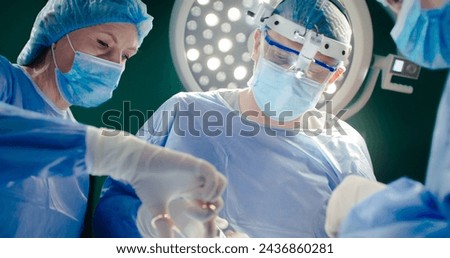 Operation, doctors dressed up in special clothes. Male doctor in binoculars. African American in special mask and gloves asssists operation. Medical workers at background of operating lamp.