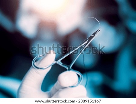 An operator holding half circle cutting needle and suture with hemostatic forceps with hand in front of of blurry operators under the lights in operating room.
