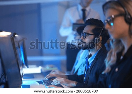 Operators wearing headsets work at computers in a dark modern call center monitoring data on glowing screens at night.