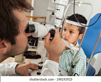 Ophthalmologist In Exam Room With Little Boy Sitting In Chair Looking Into Eye Test Machine