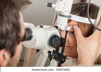 Ophthalmologist In Exam Room With Mature Woman Sitting In Chair Looking Into Eye Test Machine