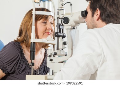 Ophthalmologist In Exam Room With Mature Woman Sitting In Chair Looking Into Eye Test Machine