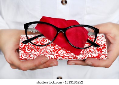 Ophthalmologist holding eyeglasses for a try out. Optometrist hands holding new black plastic eyeglasses red microfiber on white-red case, at optician shop.Close-up view focused on glasses. 