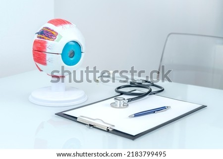 OPHTHALMOLOGIST'S DESK IN THE OFFICE WITH HUMAN EYE ANATOMY MODEL, STETHOSCOPE, PEN AND CLIPBOARD.
