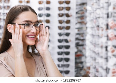 Ophthalmology And Vision Correction Concept. Portrait of smiling young female client wearing new glasses, choosing frame, standing near rack and showcase with eyewear. Woman trying on spectacles - Powered by Shutterstock