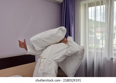 Opposite the large window stands a man in a white, terry-cloth robe, holding a pillow in his hand. The man dodges a white pillow flying at him. The concept of adult games, adults are children too. - Shutterstock ID 2180529191