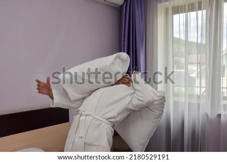 Opposite the large window stands a man in a white, terry-cloth robe, holding a pillow in his hand. The man dodges a white pillow flying at him. The concept of adult games, adults are children too.