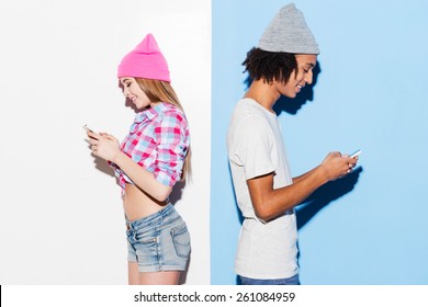 Opposites attract. Funky young couple holding mobile phones and standing back to back while standing against colorful background