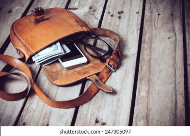 Optical Glasses, Money And Smartphone In An Open Leather Hipster's Bag On A Wooden Board Background.