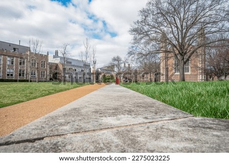 Optical illusion. Photo taken on a cement ledge but looks like it's the same level as the sidewalk. Pathway with green grass on both sides. Old buildings. Blue sky with white clouds.