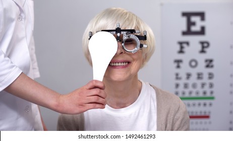 Optician Closing One Eye To Smiling Elderly Woman In Phoropter, Perfect Vision