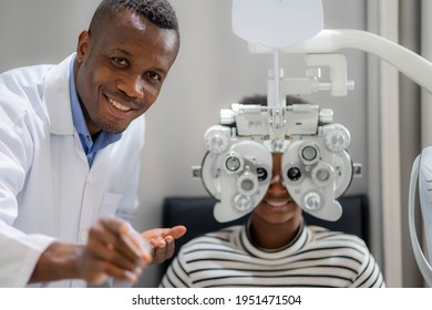 Optician Doing Optometry Eye Exam For Black African Teen Girl Patient. Male Optometrist With Phoropter While Examining Patient.