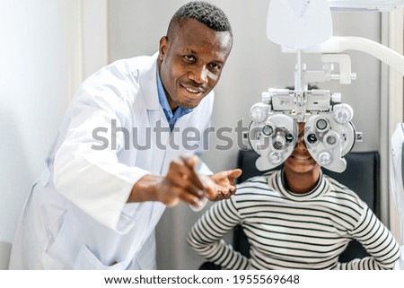 Optician doing optometry eye exam for black african american teen girl patient. Male optometrist with phoropter while examining patient.