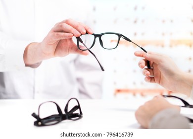 Optician Giving New Glasses To Customer For Testing And Trying. Eye Doctor With Client Comparing Spectacles And Choosing Lenses In Store.