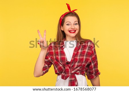 Optimism and success. Portrait of happy winner, pinup girl in checkered shirt and headband showing v sign or peace, victory gesture. retro vintage 50's style, studio shot isolated on yellow background