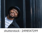 Optimistic adult black hipster male in hat and glasses peeking out of pillar of modern urban building and looking away with curiosity