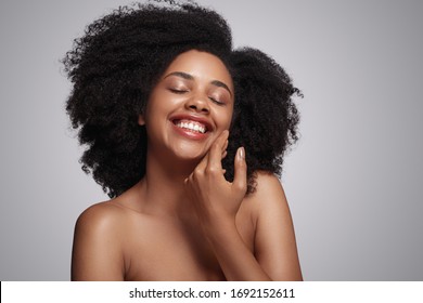 Optimistische Afroamerikanerin mit lockigen Haaren, die mit geschlossenen Augen lächelt und die perfekte Haut der Wange auf grauem Hintergrund berührt