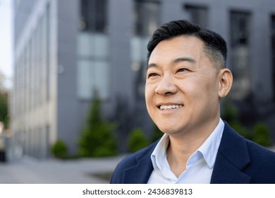 An optimistic Asian business professional smiling confidently in an urban setting, with a blurred modern building background. - Powered by Shutterstock