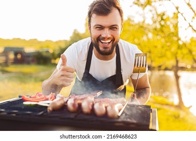 Optimistic bearded male chef with spatula looking at camera with smile and gesturing thumb up, while grilling sausages and vegetables during picnic on lake shore in summer weekend - Shutterstock ID 2169468623