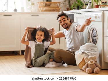 Optimistic Black Family Father And Happy Son In Casual Clothes Smiling And Covering Themselves With Towel While Sitting On Floor And Doing Laundry In Kitchen At Home