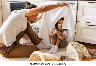 Optimistic Black Family Father And Happy Son In Casual Clothes Smiling And Covering Themselves With Towel While Sitting On Floor And Doing Laundry In Kitchen At Home