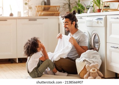 Optimistic Black Family Father And Happy Son In Casual Clothes Smiling And Covering Themselves With Towel While Sitting On Floor And Doing Laundry In Kitchen At Home