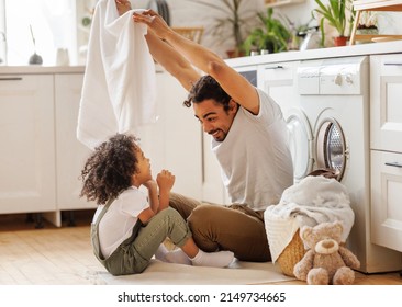 Optimistic Black Family Father And Happy Son In Casual Clothes Smiling And Covering Themselves With Towel While Sitting On Floor And Doing Laundry In Kitchen At Home