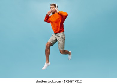 Optimistic Brown Haired Man With Beard In Sweatshirt, Shorts And Sneakers Holding White Headphones And Jumping On Isolated Background..