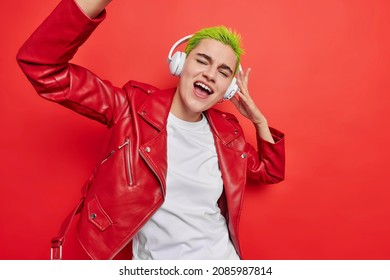 Optimistic carefree fashionable woman with green short hair sings song along shakes arms enjoys favorite music in headphones wears stylish leather jacket isolated over vivid red background has fun - Powered by Shutterstock