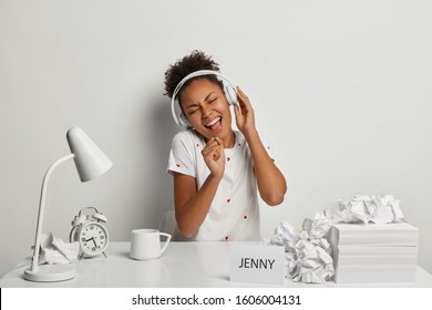Optimistic dark skinned woman has fun at workplace, listens music in stereo headphones and sings song loudly, wears casual white t shirt, sits at desktop with much papers, alarmclock, cup of drink