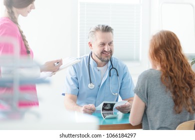 Optimistic Doctor Explaining Ultrasound Image To His Patient And Nurse Taking Notes