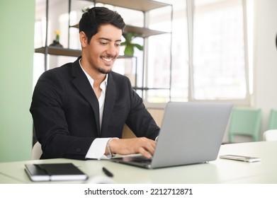 Optimistic ethnic man in formal business wear using laptop sitting on the workplace in modern open space office, young hispanic male employee replying emails, working on a new project - Shutterstock ID 2212712871