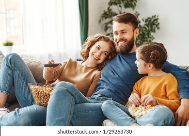 Optimistic Family In Casual Wear Sitting On Sofa In Light Room And Laughing While Watching TV And Eating Tasty Popcorn During Sunny Day At Home
