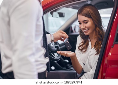 Optimistic Female Client Sitting In Brand New Car And Taking Keys From Crop Dealer In Dealership