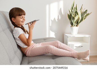 Optimistic Funny Little Cute Dark Haired Female Child Wearing White Casual T Shirt Using Phone While Sitting On Comfortable Couch, Recording Message Or Use Voice Assistant.