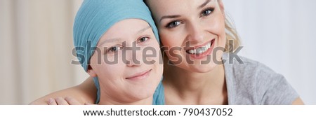Optimistic girl with cancer wearing blue scarf with smiling sister supporting her