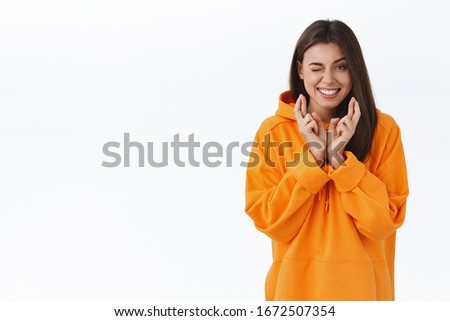 Optimistic good-looking modern girl in orange hoodie peeking at camera while cross fingers good luck and making wish, smiling hopeful, praying for dream come true, believe in miracles