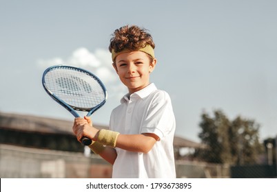 Optimistic little boy with curly hair holding racket and looking at camera while preparing for hit during training in tennis club in summer day - Powered by Shutterstock